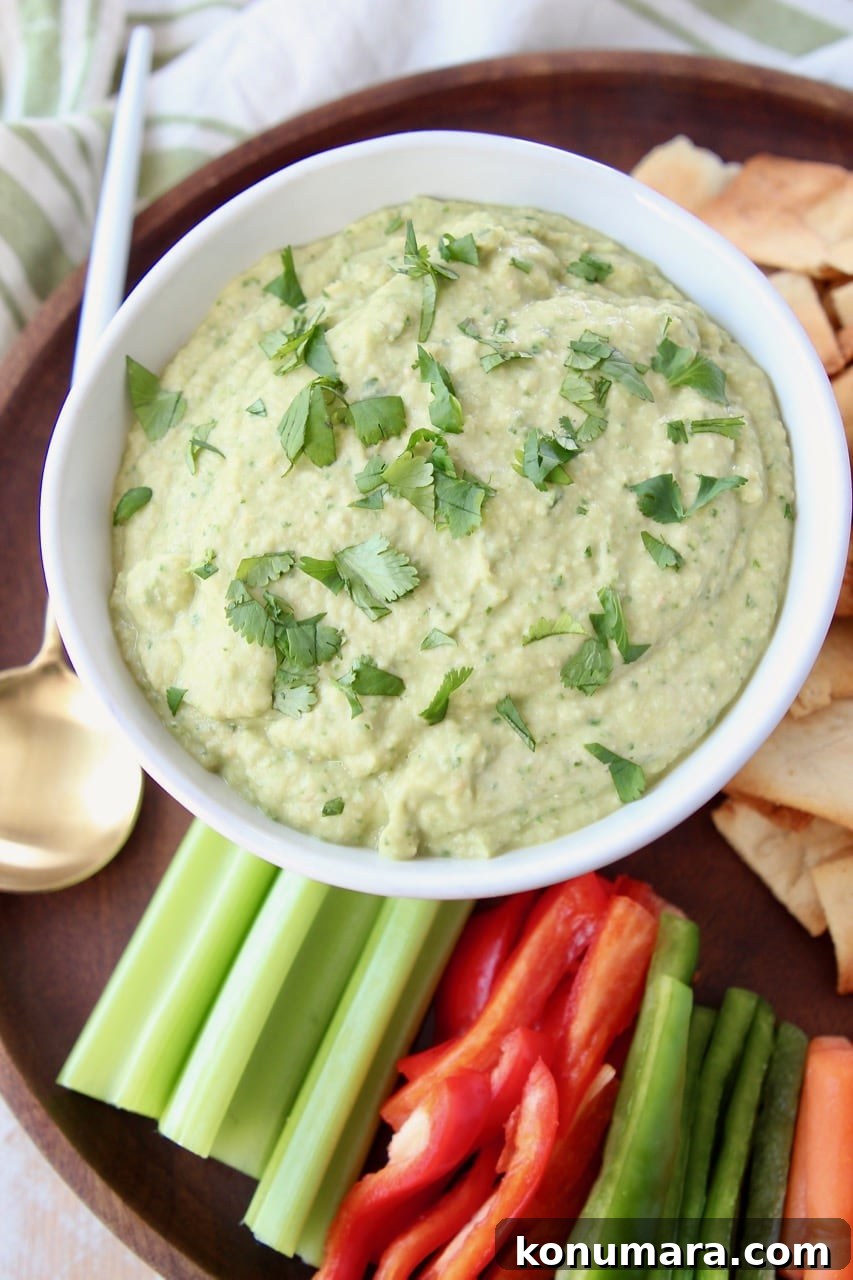 Spinach artichoke hummus in white bowl on wood serving tray with fresh celery, carrots and bell peppers
