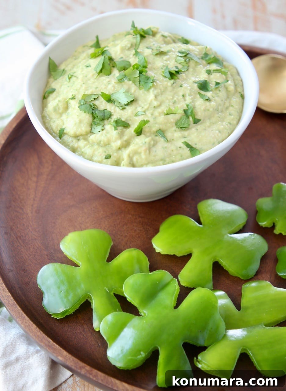 Spinach artichoke hummus in a white bowl on a wood serving tray with shamrock shaped green bell peppers
