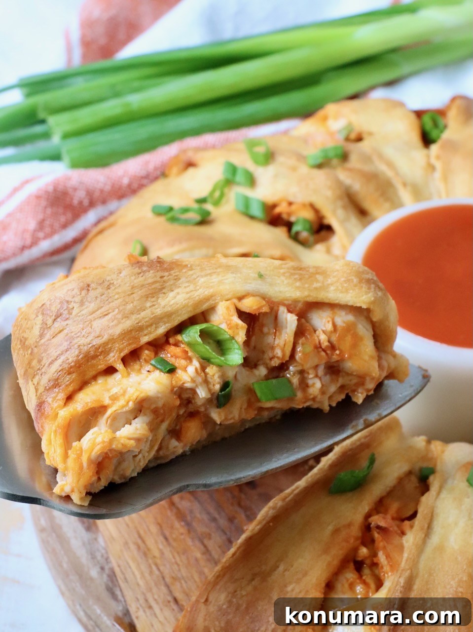 slice of buffalo chicken crescent ring on a spatula with green onions and buffalo sauce in a bowl on the side