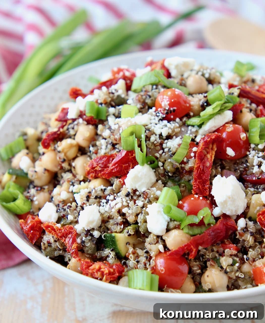 Cooked quinoa, sun dried tomatoes, chickpeas and feta cheese in white bowl topped with green onions