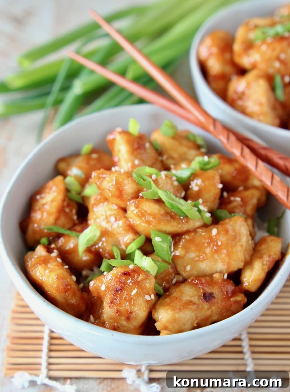 Orange chicken in bowl topped with sliced scallions and sesame seeds, with wood chopsticks on the side of the bowl