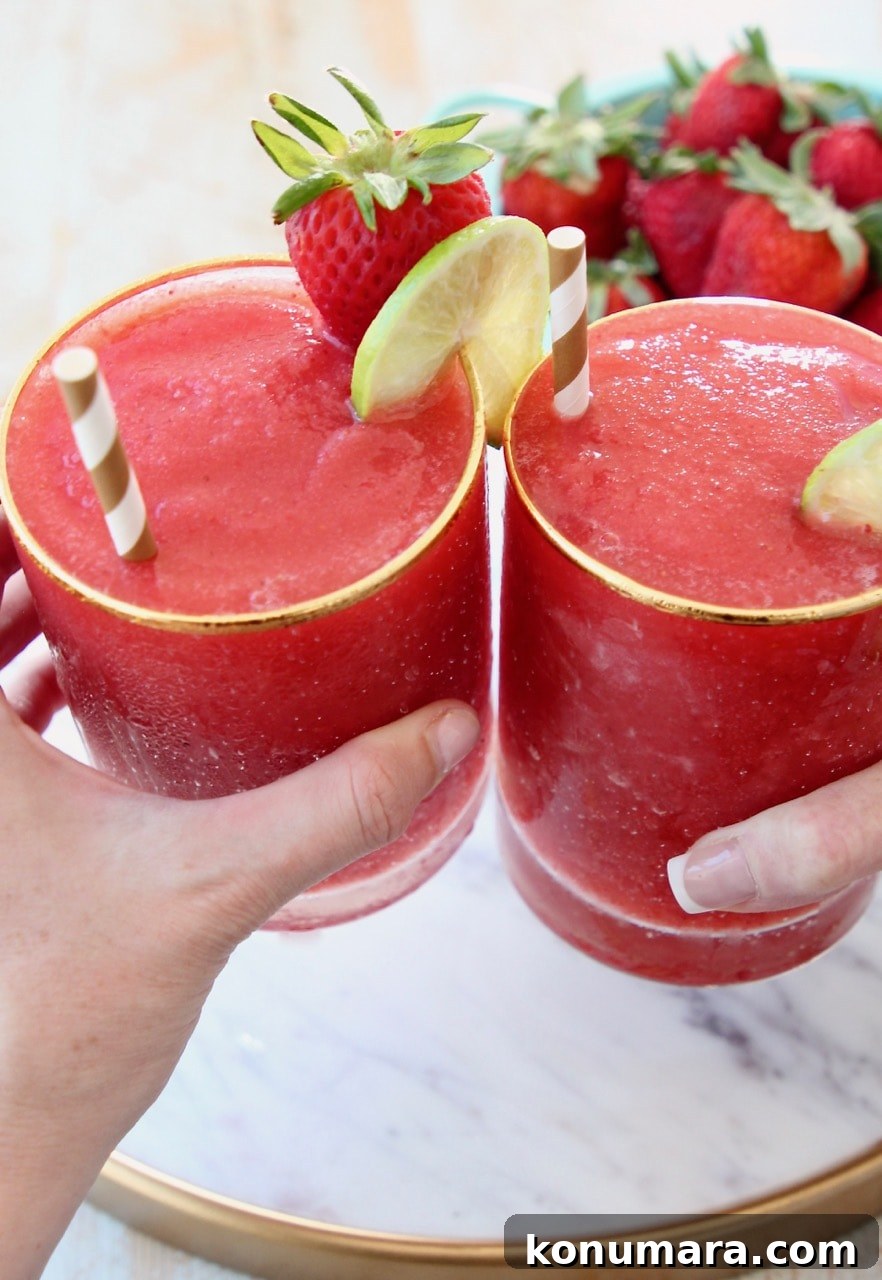 Strawberry Daiquiri Recipe with Basil-Infused Simple Syrup 3 Two hands holding glasses of strawberry daiquiris with gold and white straws and lime wedges on the side