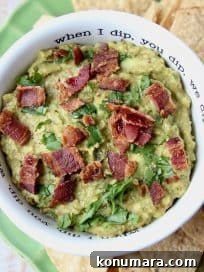 Guacamole in white bowl topped with cooked bacon, with tortilla chips on the side