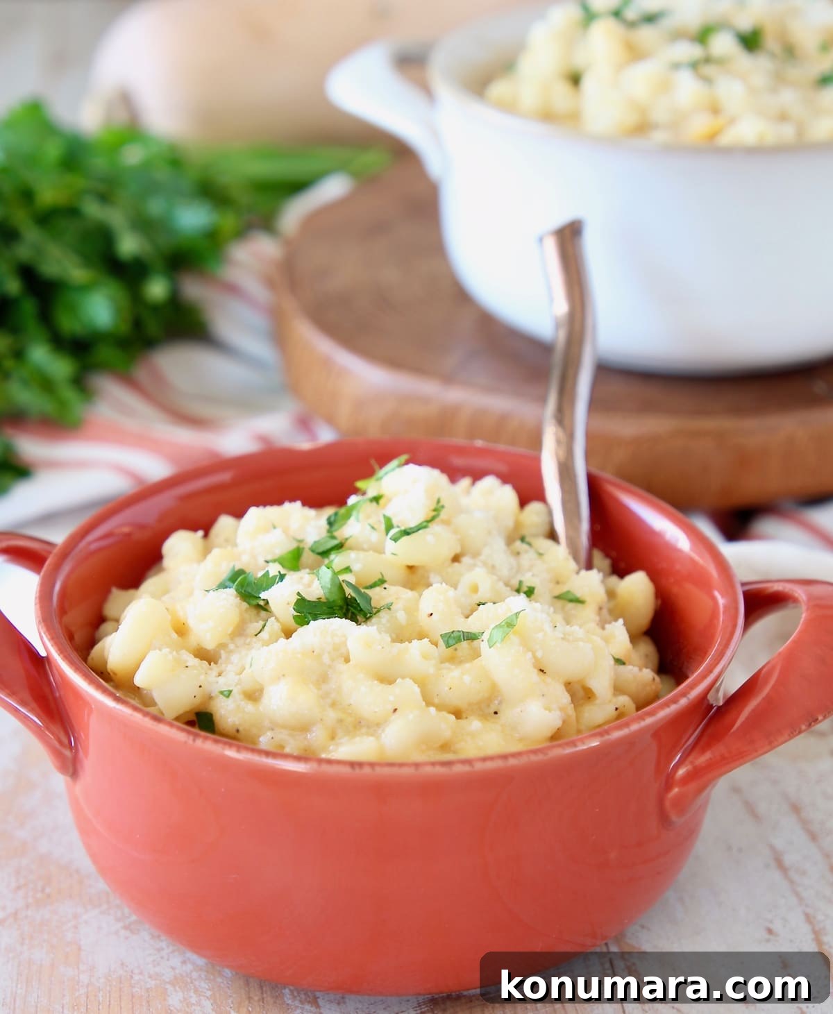 Butternut Squash mac and cheese in orange crock with gold spoon and fresh parsley with white bowl of mac and cheese in the background