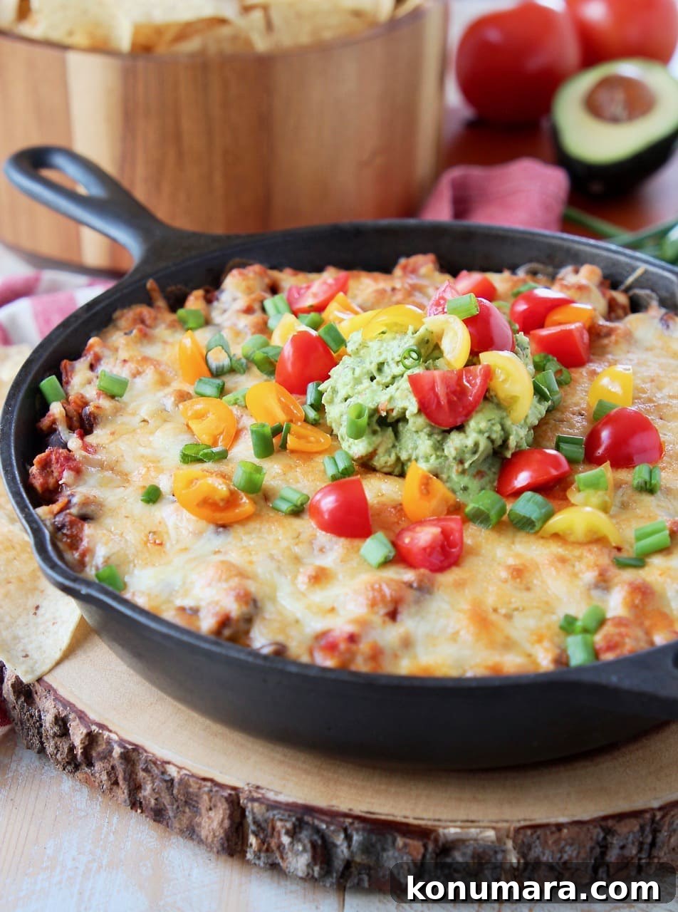 Baked taco dip in a cast iron skillet, sitting on a wood cutting board with a bowl of chips in the background