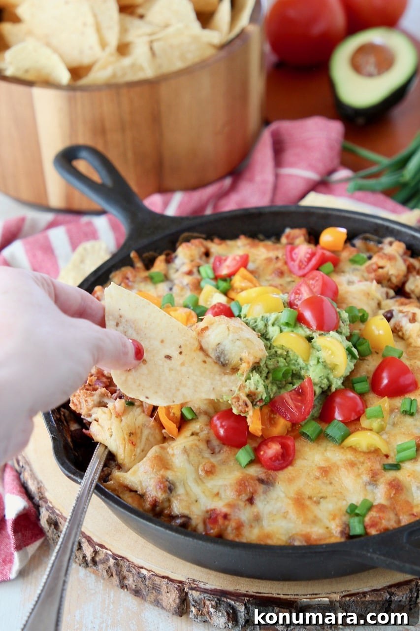 Hand dipping chip into cheesy baked taco dip topped with guacamole and diced tomatoes