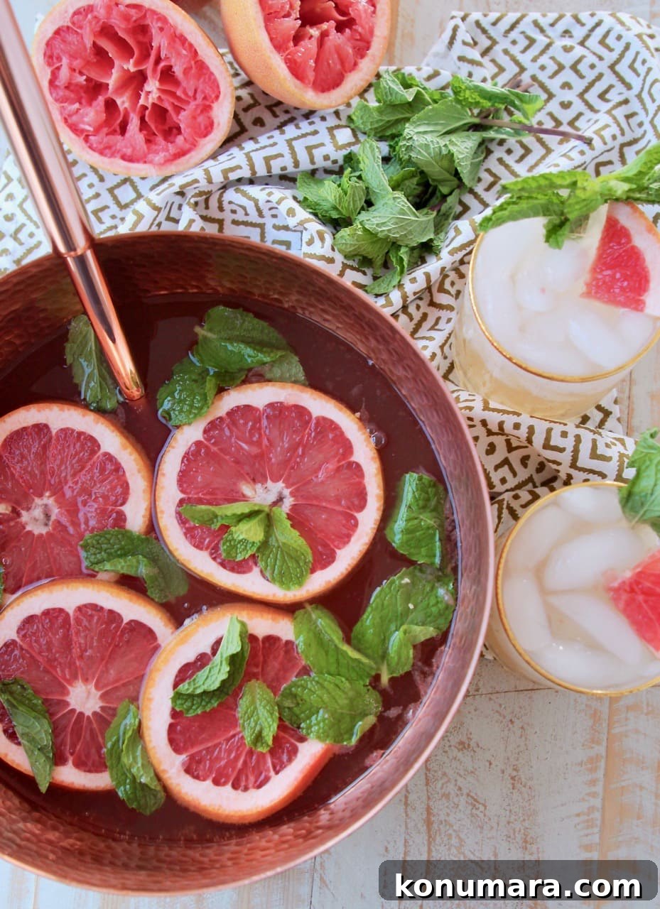 Grapefruit and Ginger Spiced Rum Punch Recipe 2 Copper serving bowl filled with spiced rum punch, topped with slices of grapefruit and mint leaves, with two glasses of rum punch on the side sitting on a patterned towel with fresh mint leaves
