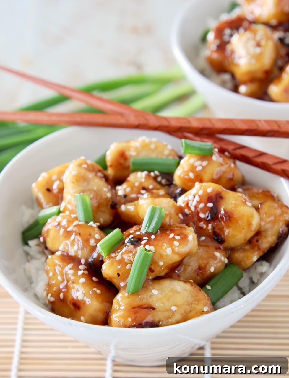 Gluten-Free Chinese Honey Chicken Recipe 7 Chinese honey chicken pieces in white bowl with white rice, garnished with green onions and sesame seeds with brown chopsticks on the side of the bowl and whole green onions in the background