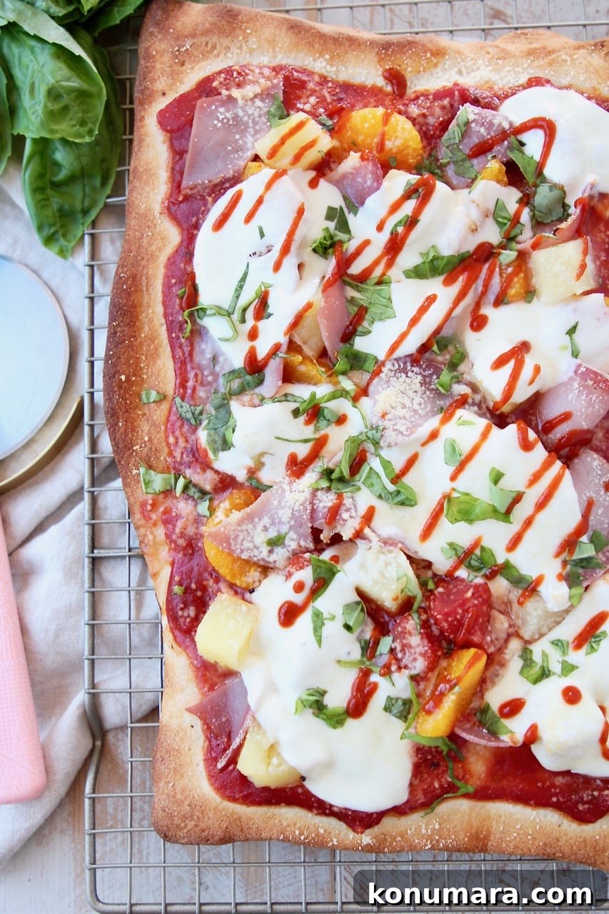 Hawaiian pizza on wire rack with sriracha drizzled on top and pink pizza cutter on the side