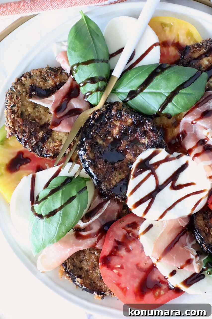 A vibrant Caprese salad with sliced tomatoes, mozzarella, crispy eggplant, prosciutto, fresh basil, and balsamic glaze drizzle on a white plate.