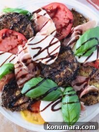 A vibrant Caprese salad with sliced tomatoes, mozzarella, crispy eggplant, prosciutto, fresh basil, and balsamic glaze drizzle on a white plate.