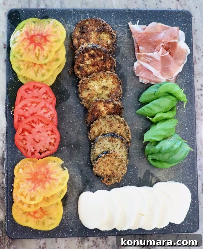 sliced heirloom tomatoes, crispy fried eggplant slices, sliced mozzarella cheese, basil leaves, and slices of prosciutto on a black cutting board
