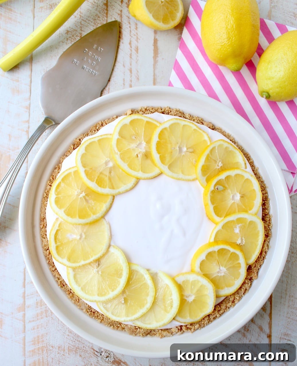 A creamy lemon pie topped with lemon slices sits on a wooden table. Beside it are whole lemons, a striped napkin, and a pie server.
