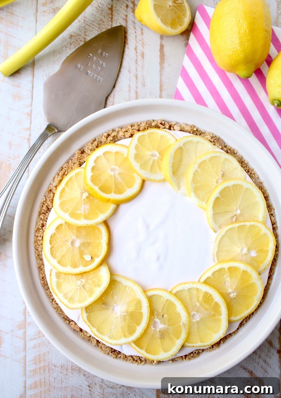A creamy lemonade pie topped with lemon slices sits on a wooden table. Beside it are whole lemons, a striped napkin, and a pie server.