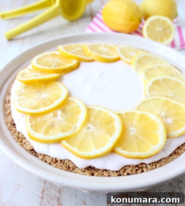 A creamy lemon pie topped with lemon slices sits on a wooden table. Beside it are whole lemons, a striped napkin, and a pie server.