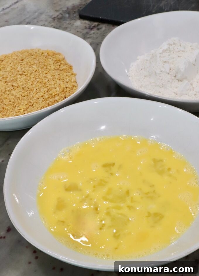three bowls on a countertop, one with whisked eggs, one with flour, and one with crushed cornflakes