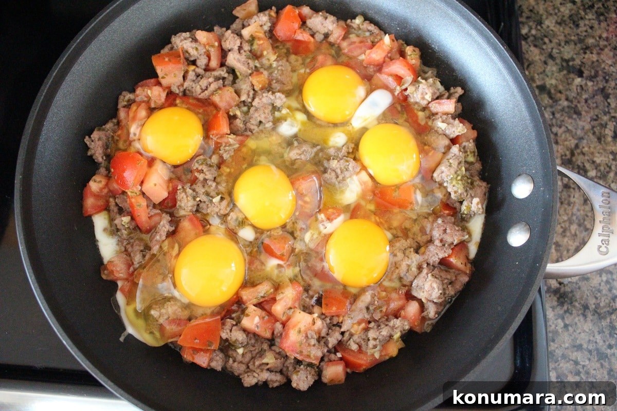 raw eggs on top of cooked sausage with diced tomatoes in a skillet