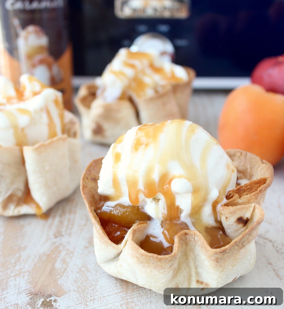 crispy taco shell cups filled with slow cooked fruit, topped with a scoop of ice cream and caramel syrup on a white wood board in front of a blue crock pot