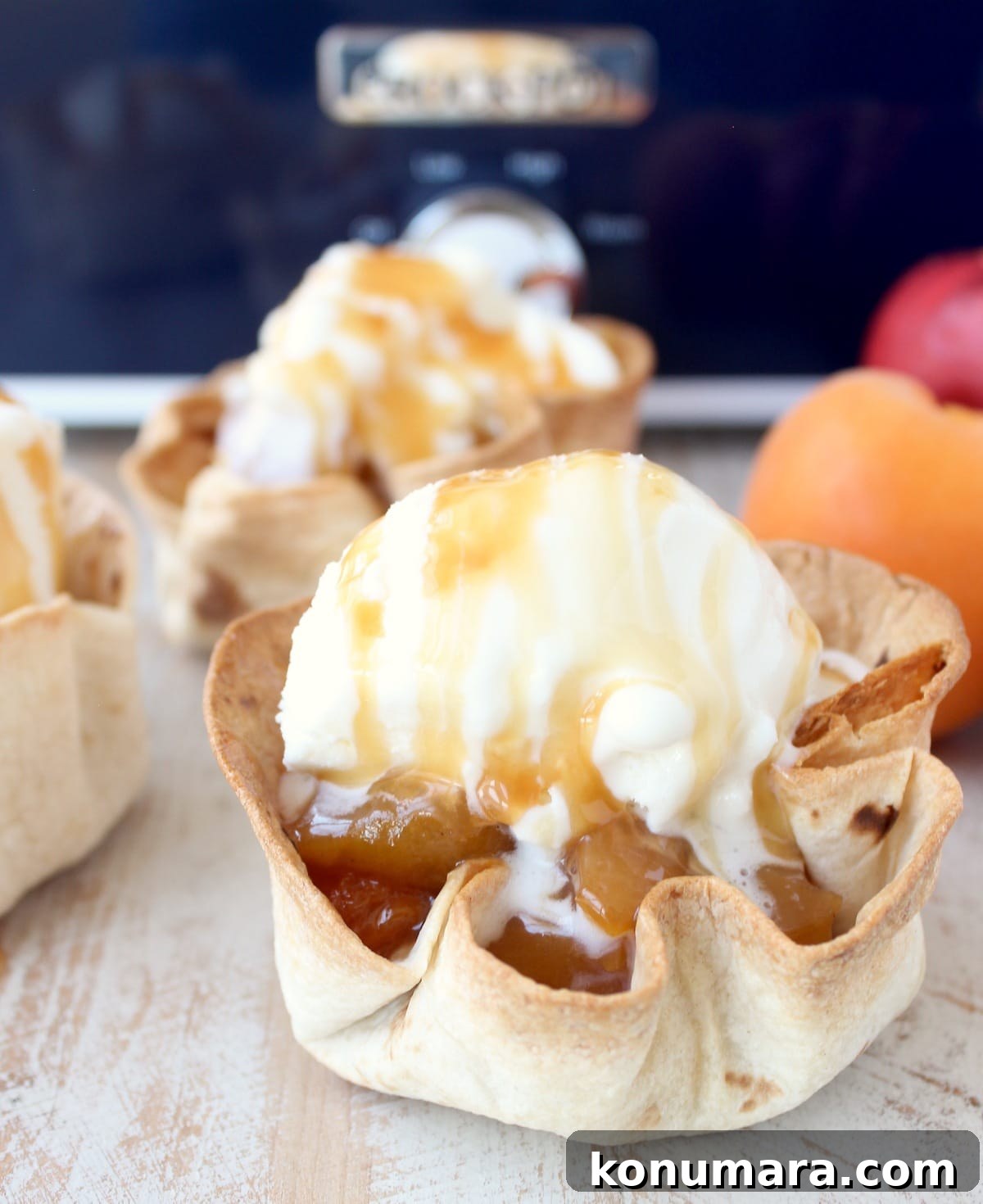 crispy taco shell cups filled with slow cooked fruit, topped with a scoop of ice cream and caramel syrup on a white wood board in front of a blue crock pot