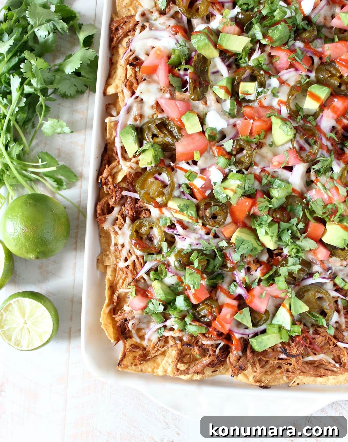 Loaded Pulled Pork Nachos Recipe with Tangy BBQ Sauce 2 A white dish filled with tortilla chips topped with pulled pork, cheese, onions, tomatoes, and avocado on a wooden surface next to limes and cilantro.