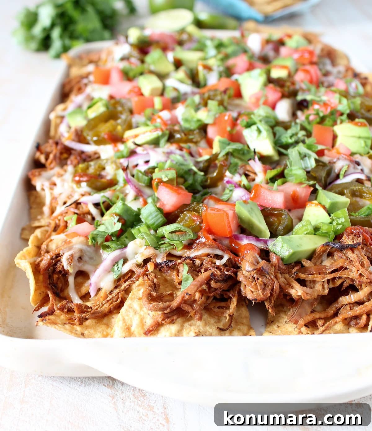 Loaded Pulled Pork Nachos Recipe with Tangy BBQ Sauce 6 A white dish filled with tortilla chips topped with pulled pork, cheese, onions, tomatoes, and avocado.
