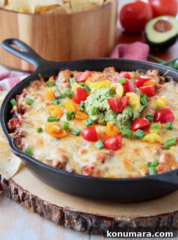 Holiday Gifts Foodies Will Love 3 Baked taco dip in a cast iron skillet, sitting on a wood cutting board with a bowl of chips in the background, sitting on a red and white striped towel