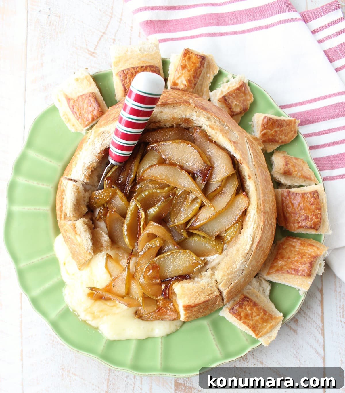 Baked brie in bread bowl with caramelized pears and honey on green plate with red and white cheese spreader and towel