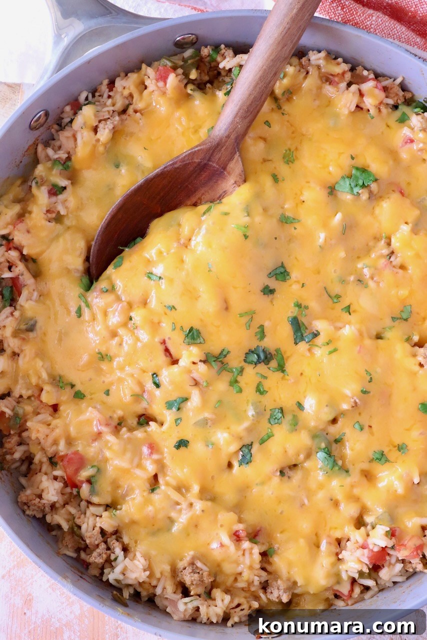 A skillet filled with cheesy Mexican rice, topped with melted golden cheese and garnish of green herbs. A wooden spoon is scooping from the side.