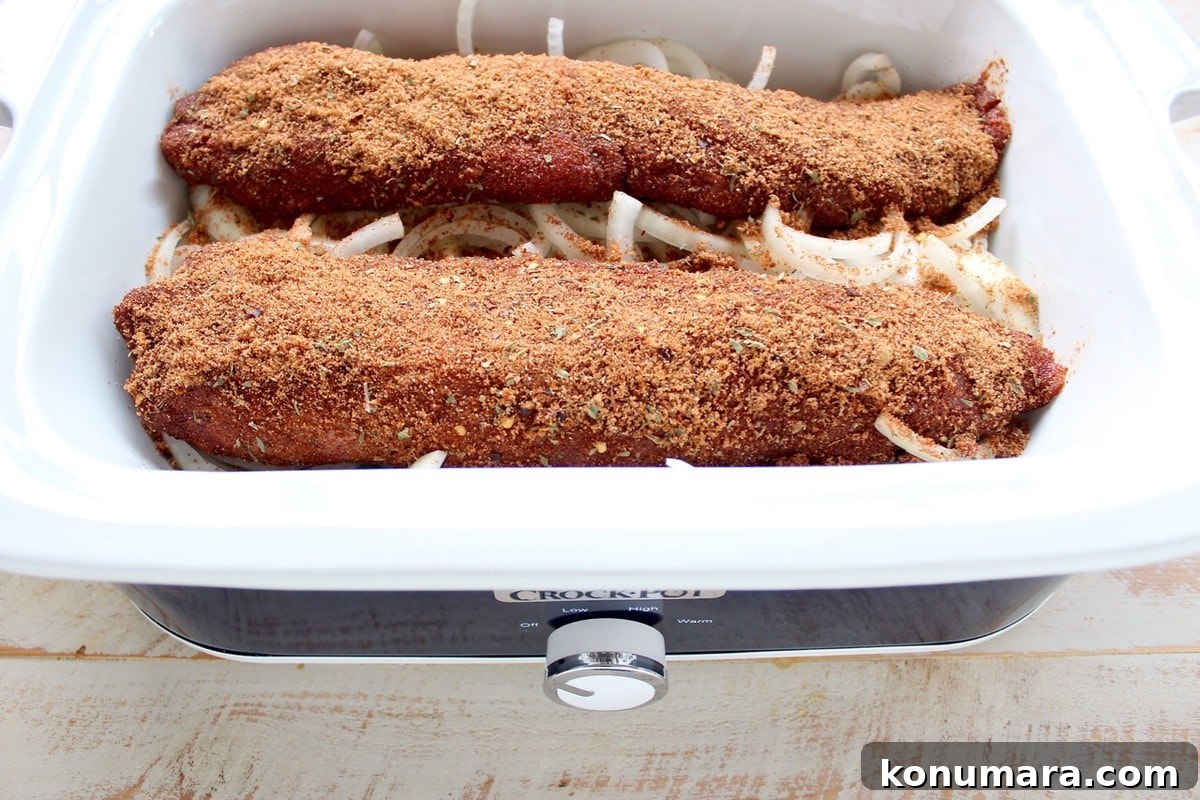 Pork tenderloins coated in a dry rub set on top of onions in a slow cooker.
