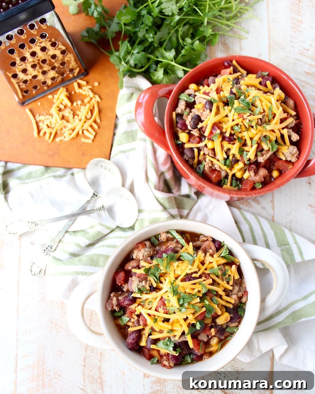 Two bowls filled with chili and shredded cheese on a wooden surface surrounded by a green and white napkin, spoons, herbs, and a cutting board with shredded cheese.