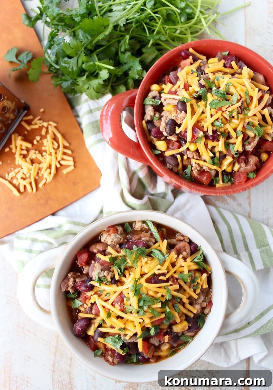 Two bowls filled with chili and shredded cheese on a wooden surface surrounded by a green and white napkin, spoons, herbs, and a cutting board with shredded cheese.