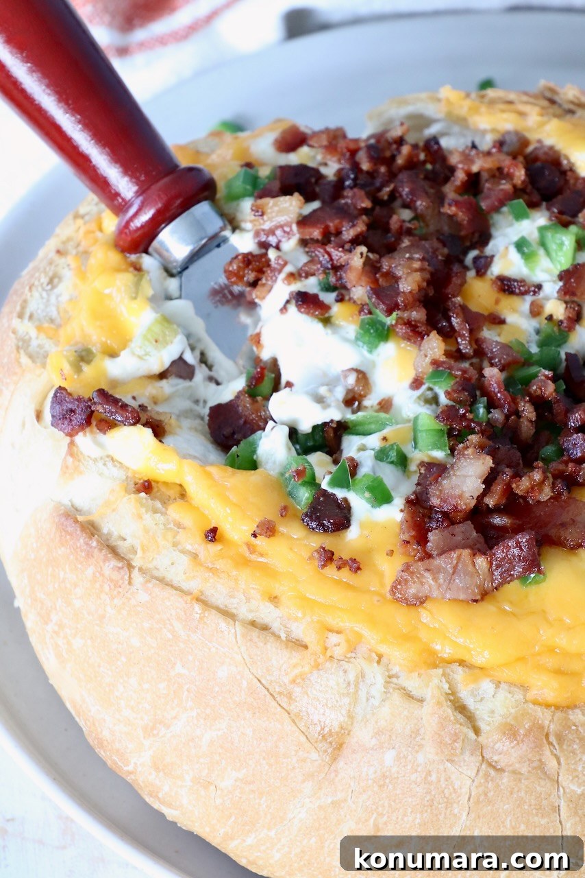 Cheesy Jalapeno Popper Bread Bowl Recipe 2 A round loaf of bread filled with melted cheese, bacon bits, and diced jalapeños. A bread knife with a wooden handle is inserted on the side.