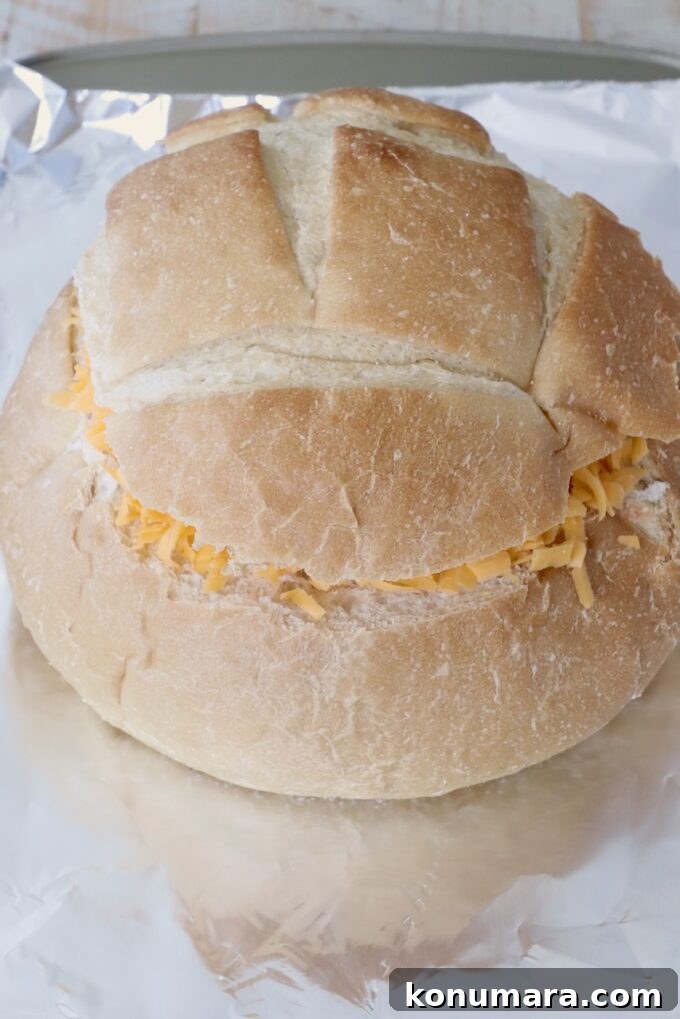 Cheesy Jalapeno Popper Bread Bowl Recipe 6 A round bread loaf with its top sliced off, filled with shredded cheese, rests on a sheet of aluminum foil on a baking sheet.