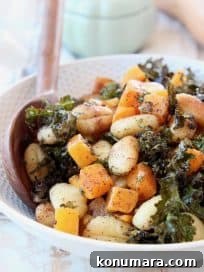 Baked gnocchi, butternut squash and kale in bowl with wooden serving spoons, whole butternut squash and green baking dish in back