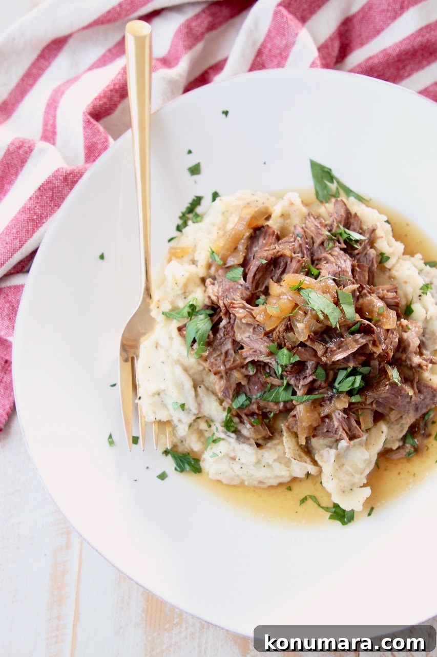 Savory French Onion Pot Roast with Caramelized Onions 13 Shredded french onion pot roast on top of mashed potatoes on a plate, topped with fresh chopped parsley with a gold fork and red and white towel