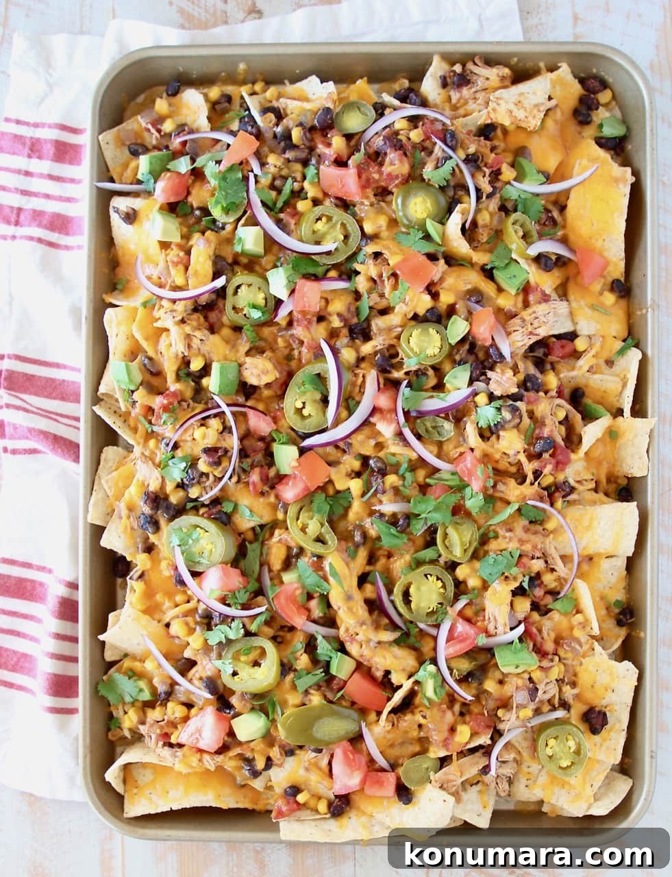 Cheesy Crock Pot Chicken Nachos Recipe 4 Sheet pan of chicken nachos topped with cheese, tomatoes, avocado, onions and jalapenos on red and white striped towel