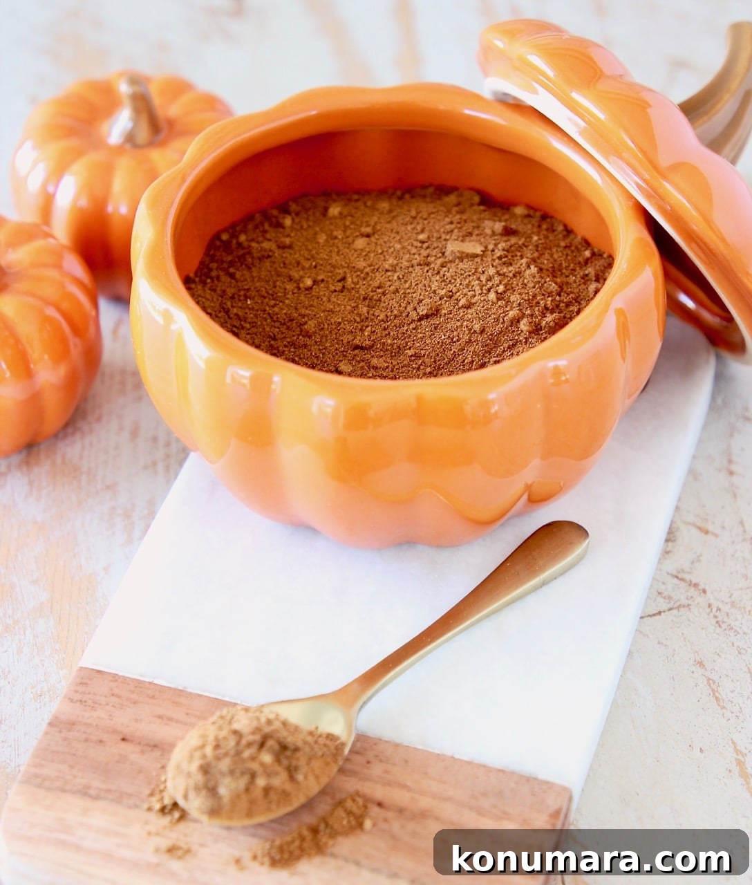 How to Make Pumpkin Pie Spice Blend 6 Pumpkin pie spice on small gold spoon on cutting board with ceramic pumpkin filled with pumpkin pie spice and two smaller orange pumpkins on the side