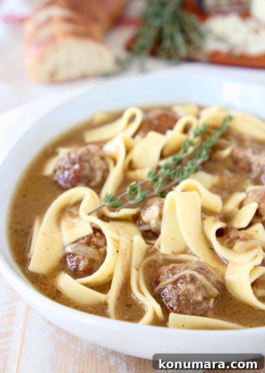 Slow Cooker French Onion Soup with Meatballs 2 Meatballs and egg noodles in french onion soup in large white bowl with sprigs of fresh thyme and a sliced baguette in the background
