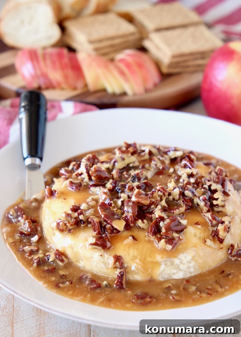 Salted Caramel Pecan Baked Brie Recipe 2 Salted Caramel and pecans on top of baked brie round on plate with cheese spreader and apples, graham crackers and sliced baguette in the background