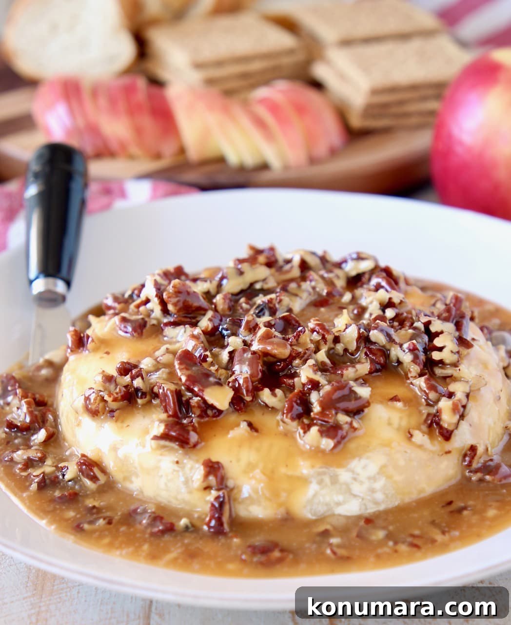 Salted Caramel Pecan Baked Brie Recipe 5 Salted Caramel and pecans on top of baked brie round on plate with cheese spreader and apples, graham crackers and sliced baguette in the background