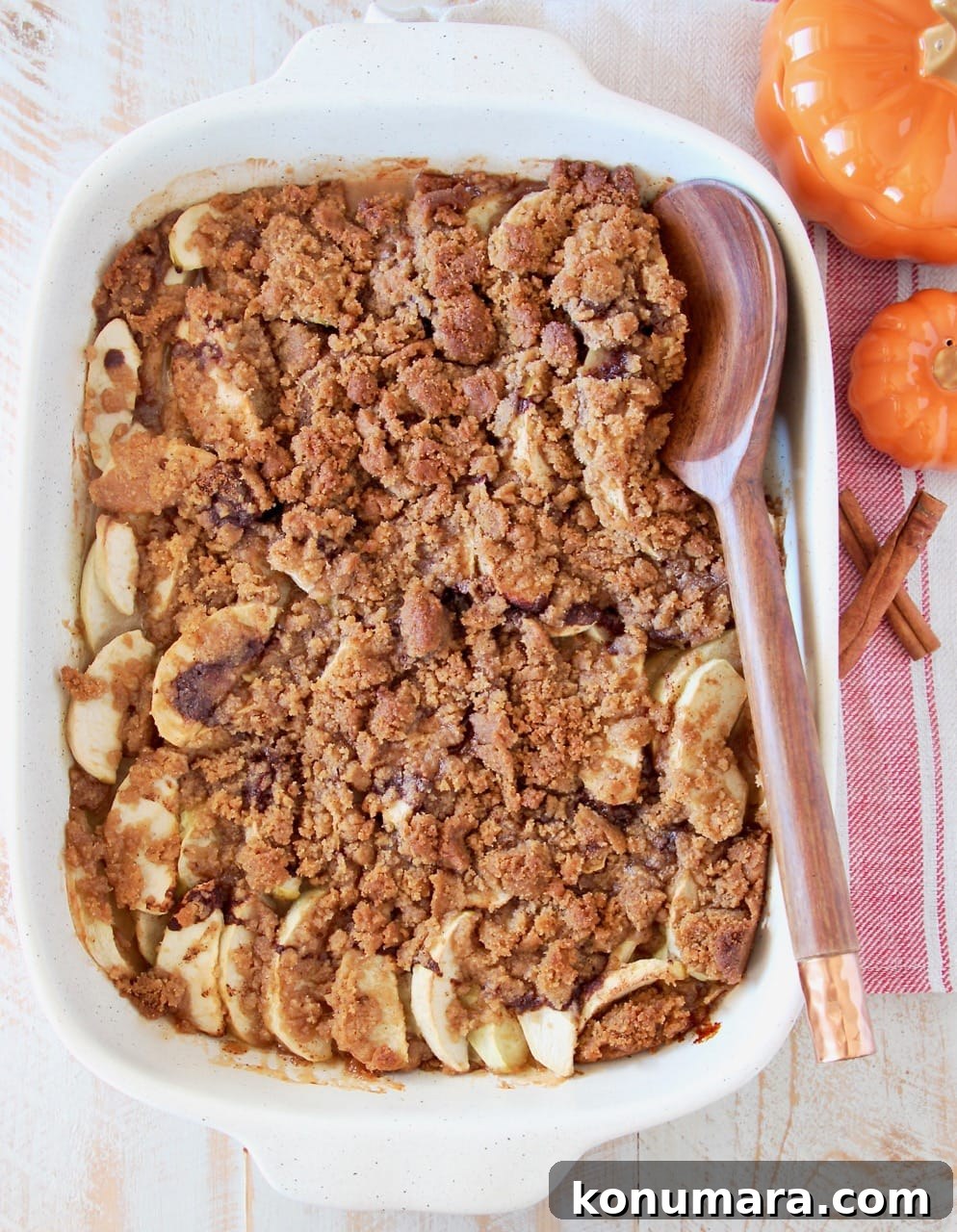 Overhead shot of apple crumble in a white casserole baking dish with a wooden spoon, sitting next to small orange pumpkins and cinnamon sticks on a red and white striped towel