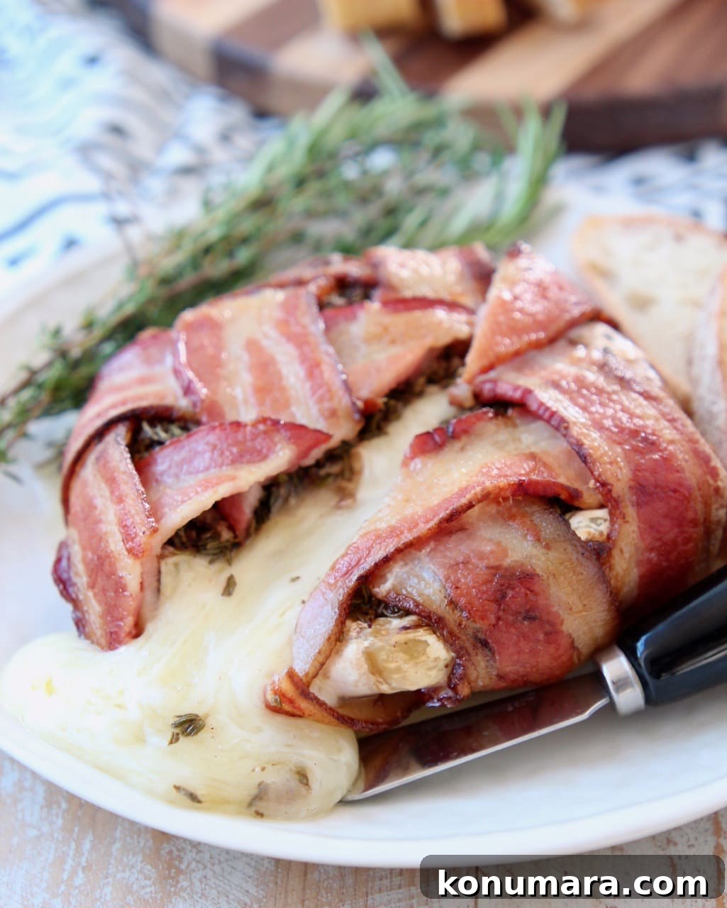 Garlic Herb Bacon-Wrapped Brie Bites 3 Baked Bacon Wrapped Brie with brie cheese pouring out onto plate with cheese spreader, fresh herbs and a sliced baguette