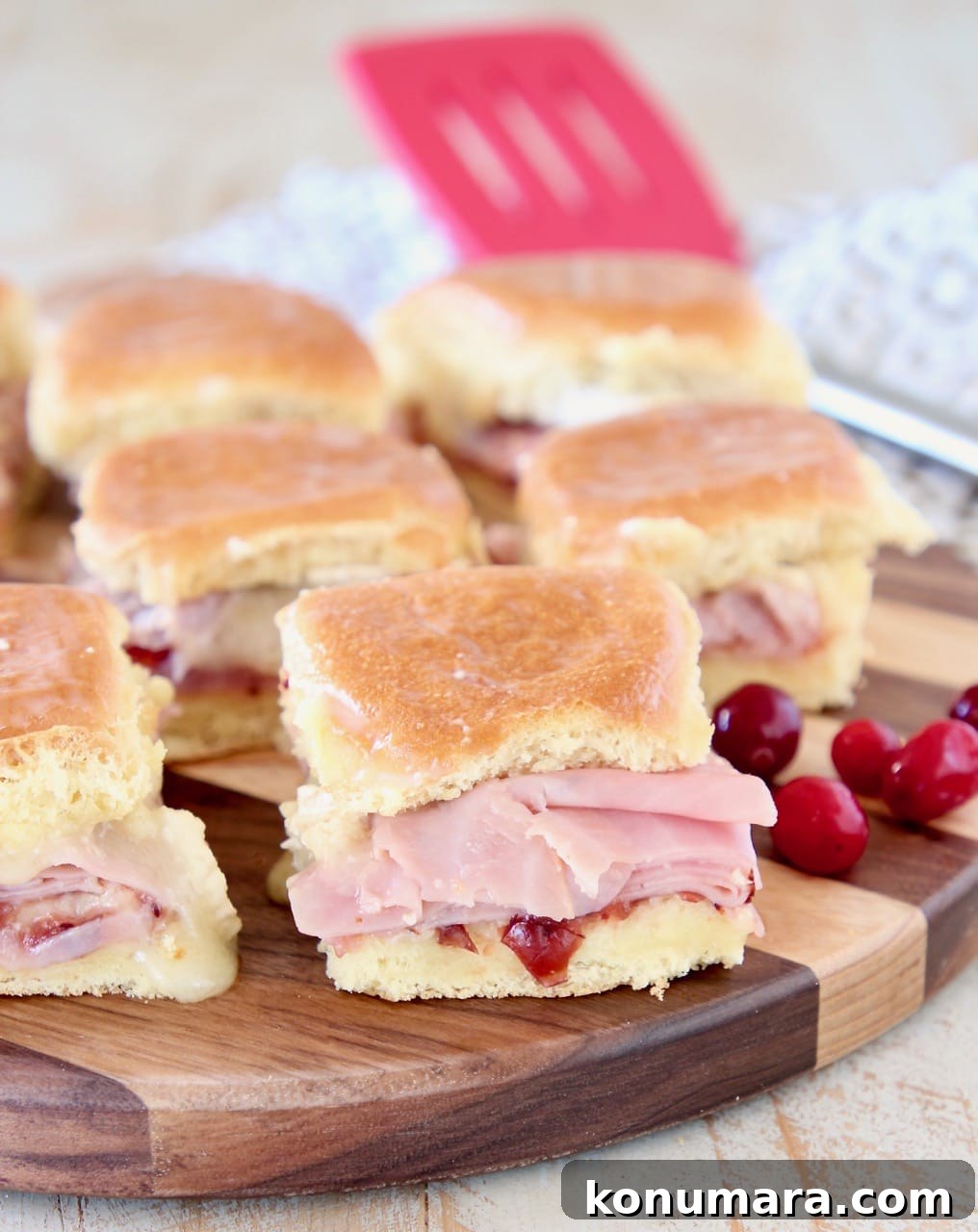 Cranberry, Brie and Ham Sliders Recipe 2 Ham sliders on Hawaiian rolls with cranberry sauce, on wood cutting board with fresh cranberries on the side and red spatula in the background