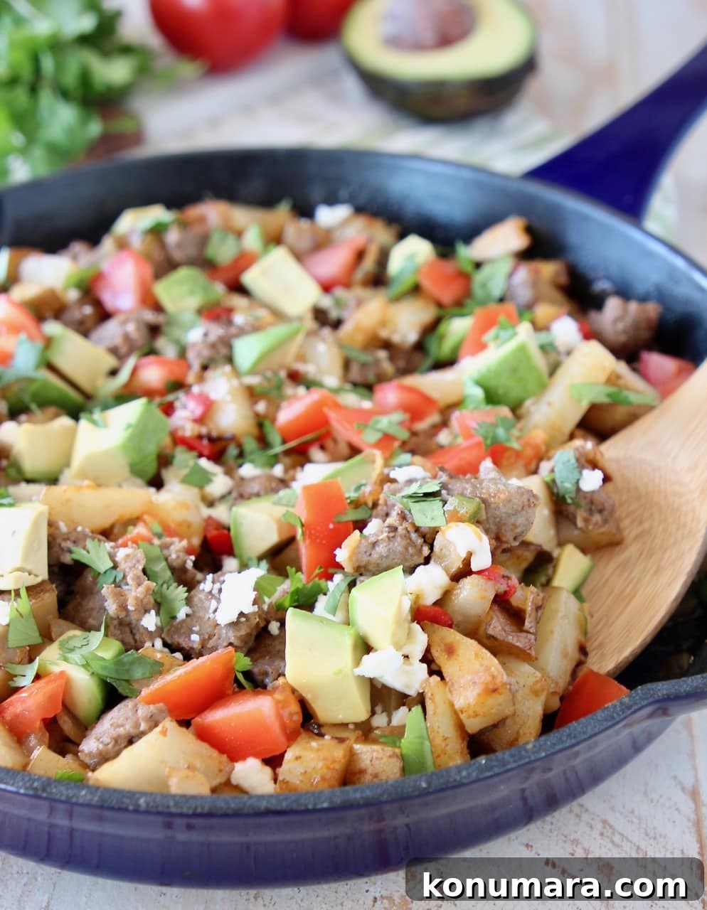 Breakfast hash in blue cast iron skillet with diced avocado, tomatoes, sausage and potatoes, with wooden spoon in skillet and fresh cilantro in background