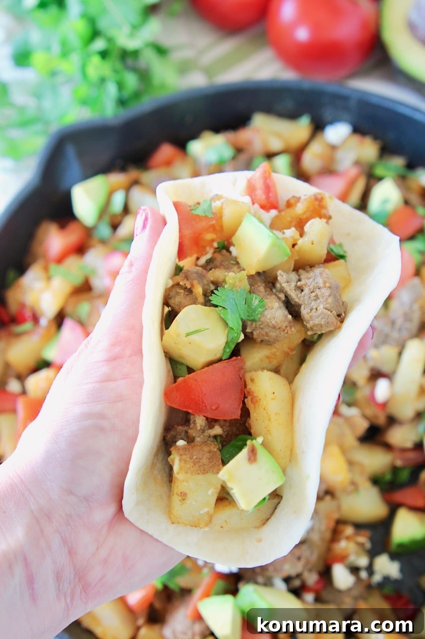 Hand holding a breakfast taco in a flour tortilla filled with diced potatoes, avocado, bell pepper, tomatoes and turkey sausage