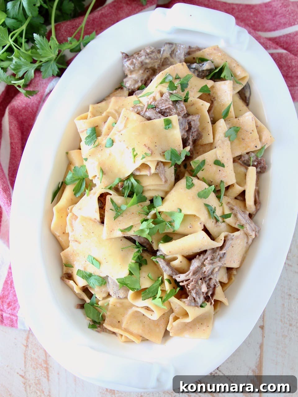 Creamy Instant Pot Beef Stroganoff 2 Instant pot beef stroganoff in a large white oval serving dish with wide egg noodles and shredded beef, topped with fresh parsley, sitting on a red and white striped towel