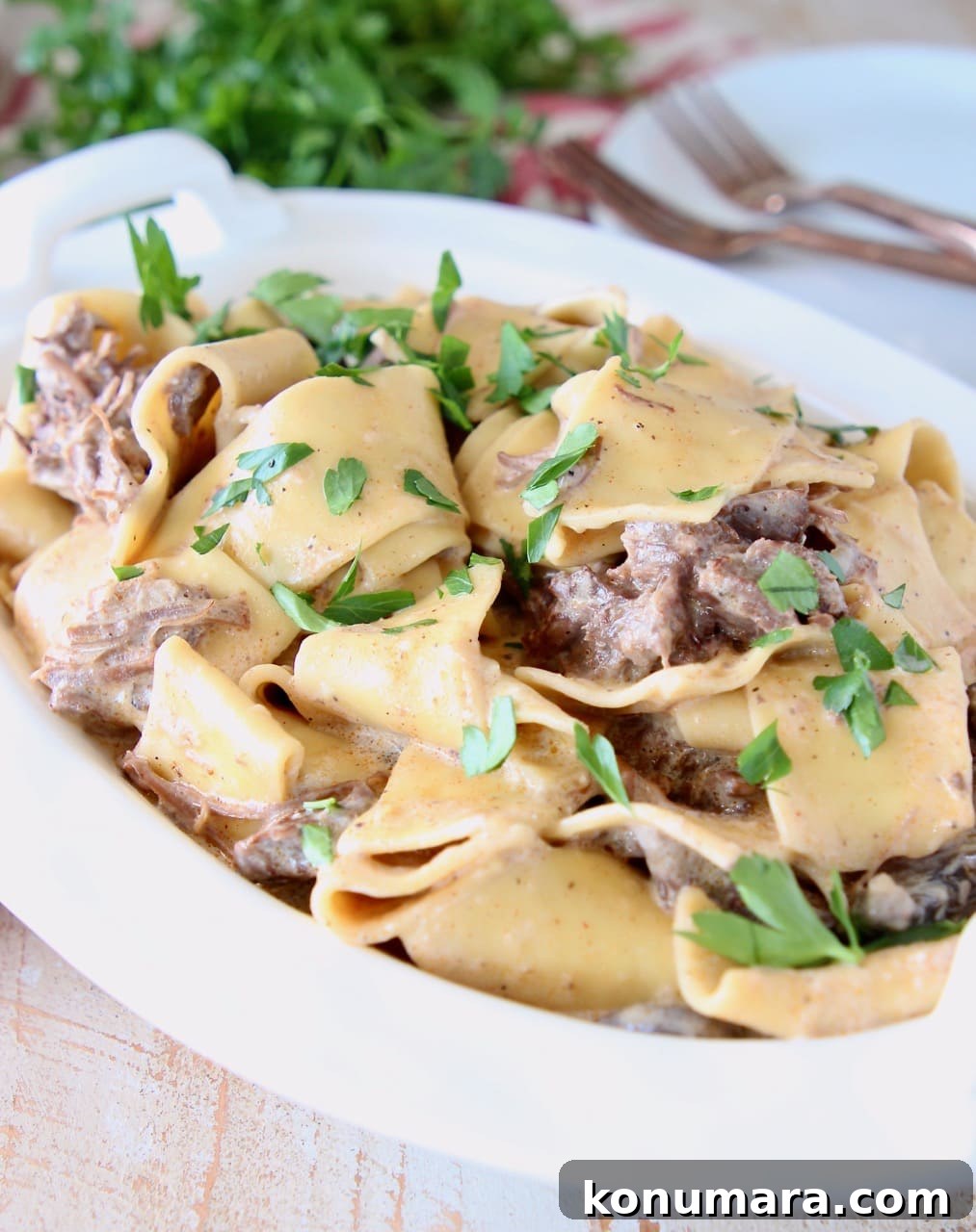 Creamy Instant Pot Beef Stroganoff 3 Beef stroganoff with wide egg noodles and fresh parsley in white serving dish, sitting on a red and white striped towel