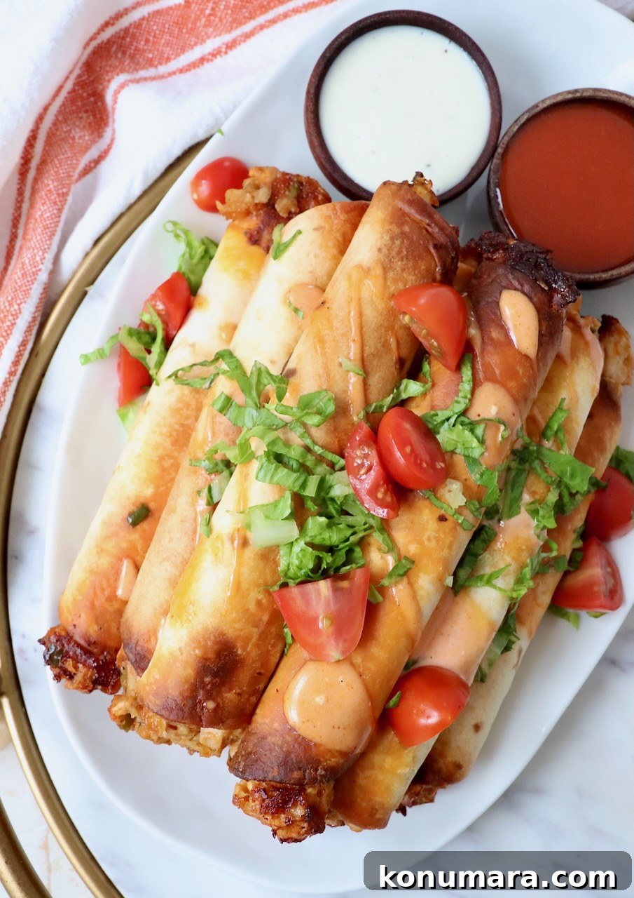 cooked taquitos stacked up on a plate topped with buffalo sauce, tomatoes, and shredded lettuce