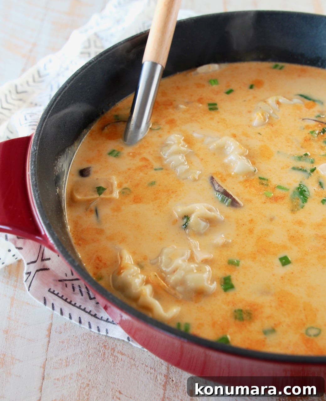 Thai coconut soup with potstickers in large red cast iron pot, garnished with cilantro and scallions, with wooden spoon handle in pot