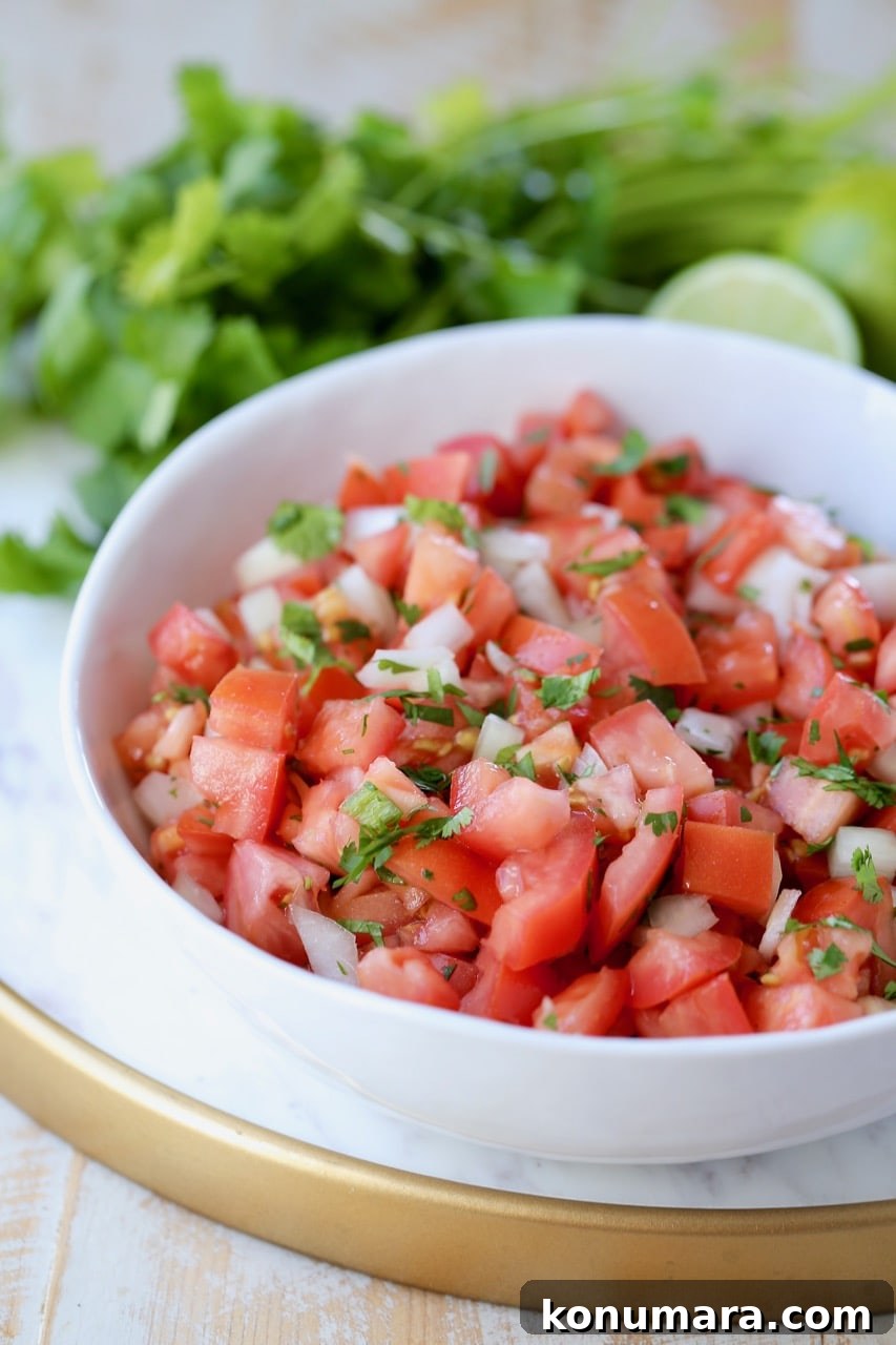 Pico de gallo in white bowl on marble serving tray, with fresh cilantro and limes in the background